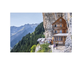 Op de afbeelding: Een houten gebouw van meerdere verdiepingen genaamd Gasthaus Aescher-Wildkirchli is direct in een steile rotswand gebouwd. De structuur heeft bruine houten muren, meerdere ramen en een terras met witte parasols dat uitkijkt over een vallei.