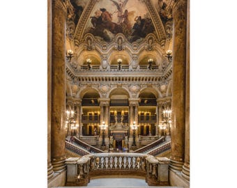 Op de afbeelding: Het grandioze interieur van het Palais Garnier toont twee massieve marmeren zuilen die een centrale trap omlijsten, sierlijke goudkleurige balkons en een gewelfd plafond met een gedetailleerde muurschildering en warme verlichting.