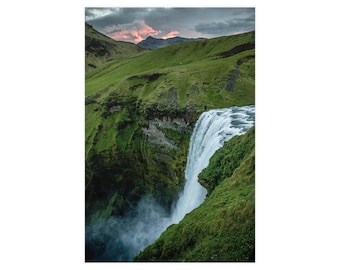 Op de afbeelding: Een grote waterval stroomt langs een steile, groene, met mos bedekte klif naar beneden in een mistig bekken. Boven de glooiende heuvels is de lucht gevuld met donkere, zware wolken die worden verlicht door een zachte roze gloed van de ondergaande zon.
