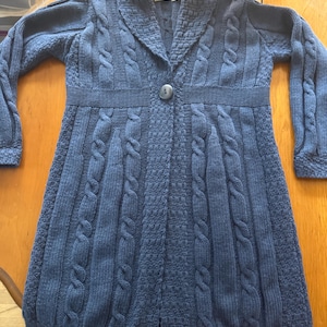 May include: A long-sleeved blue cardigan sweater featuring a shawl collar, a single central button closure, and a textured knit pattern with vertical cable knit columns. The garment includes a label at the neckline and ribbed cuffs.