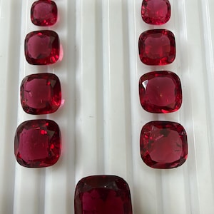 May include: Eleven faceted cushion-cut red gemstones arranged in two vertical columns of five, with one larger single gemstone centered at the bottom. Each stone displays a deep crimson hue with reflective surfaces and sharp edges.