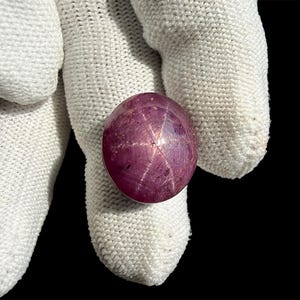 May include: A round, cabochon-cut star ruby gemstone featuring a deep magenta-pink hue and a distinct six-rayed asterism effect visible on its polished surface. The gemstone displays natural inclusions and a smooth, domed, translucent appearance.