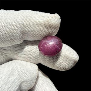 May include: A cabochon-cut star ruby gemstone with a deep magenta-red hue, displaying a distinct six-rayed asterism effect across its smooth, polished surface, held against a solid black background.