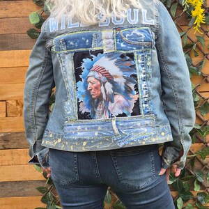 Op de afbeelding: Een lichtblauw spijkerjack met een achterpand voorzien van een grafische print van een persoon met een verentooi. Het jack heeft gerafelde denim patches, de lichtblauwe tekst WILD SOUL en gele verfspatten op de zoom.