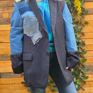 Op de afbeelding: Een marineblauw krijtstreep colbert met contrasterende blauwe spijkerstof mouwen en een opgestikte spijkerstof zak op de rechterborst. Het kledingstuk heeft twee knopen aan de voorzijde en witte decoratieve stiksels langs de revers en de mouwboorden.