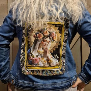 Op de afbeelding: Een blauw spijkerjack met een grote, rechthoekige geweven tapijtpatch op de achterkant. De patch toont een gedetailleerde illustratie van een figuur met een bloemenkrans en gevouwen handen, omlijst door gouden accenten en afgewerkt met rafelige randen.
