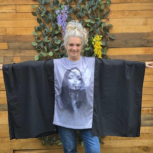 Op de afbeelding: Een zwart vest in kimono-stijl met wijde mouwen en een open voorkant, gedragen over een grijs t-shirt. Het t-shirt heeft een zwart-witte grafische print van een persoon met een kap, een kruis op het voorhoofd en een rokende sigaret.