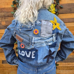 Op de afbeelding: Een blauwe spijkerjas met een patchworkontwerp op de achterkant. De jas bevat diverse stukken spijkerstof, een gele stoffen bloem, twee kleine gehaakte bloemen in oranje en paars, en een donkere stoffen ster. Het woord LOVE is opgestikt.