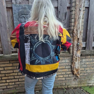 Peut inclure: Une veste en patchwork composée de panneaux de tissu rouge, jaune, noir et marron. Le dos présente un grand motif solaire circulaire en noir et gris, ainsi qu'une section de texte imprimé en noir et blanc et un ourlet à motifs.