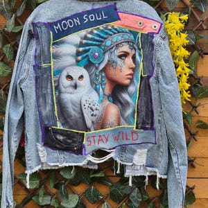Puede incluir: Una chaqueta de mezclilla azul claro con lavado ácido que presenta un gran parche rectangular en la espalda. El parche muestra a una persona con un tocado de plumas turquesa junto a un búho blanco, con el texto MOON SOUL arriba y STAY WILD en rojo.