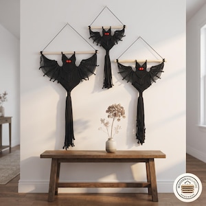May include: Three black macrame bat wall hangings suspended from light-colored wooden dowels with black string. Each bat features two bright red circular eyes and a long, fringed tail hanging downward from the center of the body.