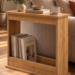 May include: A light brown wood console table with a rectangular top and a lower shelf. The top holds a small vase with greenery, a bowl, and a book titled The Kinfolk Home. The lower shelf contains a row of books, including The Kinfolk Home.