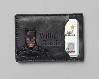 Può includere: Un portacarte in pelle nera con un rilievo della maschera di Batman che mostra il volto di una persona. Il nome William Garcia è inciso sulla parte anteriore e una patente di guida con una stella dorata è inserita nella fessura laterale.
