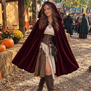 May include: A deep burgundy velvet hooded cloak worn over a cream-coloured dress with a brown lace-up corset belt. The outfit includes a small brown leather pouch, a metallic accessory, and knee-high brown boots, finished with a gold headpiece.