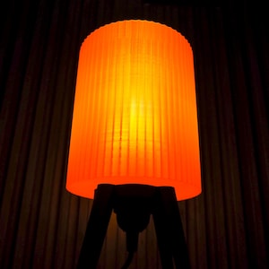 Może przedstawiać: Cylindryczna lampa z jaskrawopomarańczowym, pionowo żebrowanym kloszem, który emituje ciepłe światło. Klosz jest wsparty na ciemnej, trójnożnej podstawie. Lampa jest włączona i rozprasza światło ze środka swojej żebrowanej struktury.