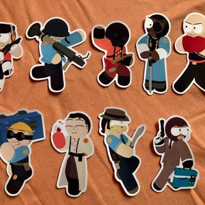 May include: A collection of nine die-cut stickers featuring stylized cartoon characters from Team Fortress 2. The figures include the Scout, Soldier, Pyro, Demoman, Heavy, Engineer with a toolbox, Medic with a medigun, Sniper with a kukri, and Spy with a briefcase.