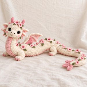 May include: A cream and light pink crocheted dragon plush toy featuring large dark eyes, small pink horns, and a long body adorned with numerous small pink flowers and green leaves along its back and tail, with a soft textured yarn finish.