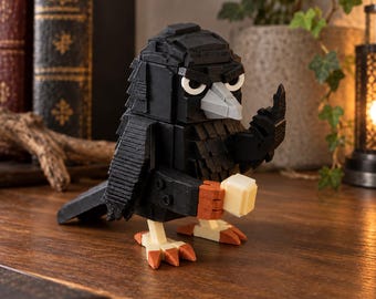 May include: A block-style crow figurine constructed from black, gray, orange, and cream-colored components. The bird features a sharp gray beak, white eyes with black pupils, and is holding a small cream-colored cube in its orange-tipped feet.