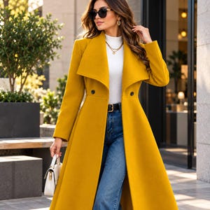 May include: A long mustard yellow wool-blend coat featuring a wide lapel, double-breasted button closure, and long sleeves. The coat is worn over a white mock-neck top and blue denim jeans, paired with a small white handbag and gold jewelry.