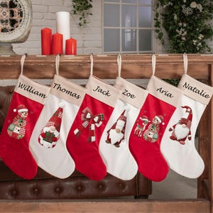 Peut inclure: Six chaussettes de Noël suspendues en rangée, chacune avec un revers beige portant un prénom brodé en écriture noire : William, Thomas, Jack, Zoe, Aria et Natalie. Les chaussettes sont rouges et blanches avec des motifs de gnomes.