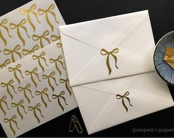 Può includere: Un foglio di adesivi in ​​foglio d'oro con un motivo a fiocco. Gli adesivi sono su uno sfondo bianco. Ci sono due buste bianche con adesivi in ​​foglio d'oro a forma di fiocco.