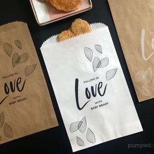 Pode incluir: Três sacos de papel, dois castanhos e um branco, com o texto "Falling in Love with Baby Brady" e ilustrações de folhas. Um saco está aberto com biscoitos dentro. Os sacos estão sobre uma superfície preta.