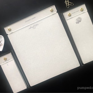 May include: Three white notepad sets with gold accents. The largest notepad has a dotted pattern and the text "pumped on paper charlotte york" printed on the top. The other two notepads have a solid white background and a single letter printed on the top.