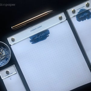 May include: Three white notepad sets with black covers and gold accents. The top notepad has a blue watercolour design with the text "charlotte york" and the brand name "pumped on paper". The middle notepad has the text "charlotte" and the brand name "pumped on paper". The bottom notepad has the letter "C" and the brand name "pumped on paper".