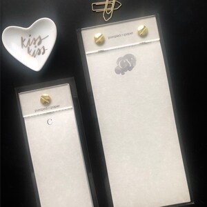 May include: Two white notepads with black text and gold accents. The notepads are in clear acrylic frames. The larger notepad has the text "pumped on paper" and the initials "CY". The smaller notepad has the text "pumped on paper" and the letter "C".