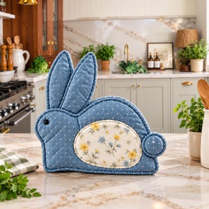 May include: A quilted fabric tea cozy shaped like a rabbit. The exterior is blue with a white polka dot pattern, featuring a black button eye. A central oval patch displays a cream background with a floral print of yellow and blue flowers.