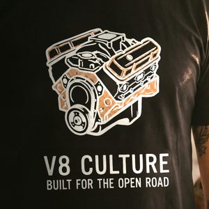May include: A black t-shirt featuring a graphic of a V8 engine in white and copper tones. Below the engine, the text reads V8 CULTURE and BUILT FOR THE OPEN ROAD in white capital letters. The design is centred on the front of the garment.