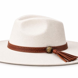 May include: A cream-coloured wide-brim fedora hat featuring a structured crown and a flat brim. A brown leather band encircles the base of the crown, accented with a circular metallic concho ornament and a dangling brown leather tassel detail.