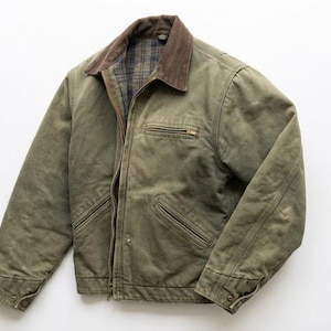 May include: An olive green canvas jacket featuring a brown corduroy collar, a front zipper closure, and a plaid interior lining. The garment includes a zippered chest pocket, two lower slanted pockets, and buttoned cuffs on the long sleeves.