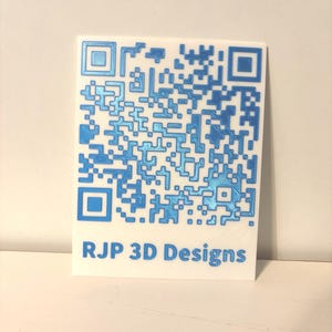 May include: A rectangular white plaque featuring a raised, metallic blue QR code pattern on the upper portion. Below the QR code, the text RJP 3D Designs is embossed in the same metallic blue colour, creating a clear contrast against the white base.
