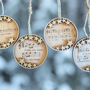 Könnte beinhalten: Vier kreisförmige Ornamente aus hellem Holz, die an Bindfäden hängen. Jedes Ornament ist mit lasergeschnittenen Sternen an den Rändern verziert und zeigt gedruckte Notenblätter mit deutschen Liedtexten wie Leise rieselt der Schnee.