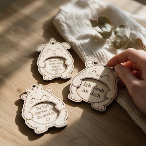 May include: Three light-coloured wooden ornaments shaped like cartoon monsters, each featuring a circular centre with engraved German text. The text reads: Du musst nicht alles alleine schaffen, Ich glaub an dich, and Du bist stärker, als du denkst.