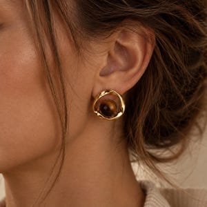 May include: A gold-toned stud earring featuring a polished, irregular circular frame that surrounds a smooth, spherical tiger's eye gemstone. The earring has a warm, metallic lustre and a dark brown, banded stone at its centre, creating a contrast.