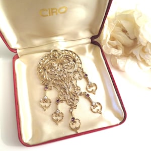 Peut inclure: Un pendentif filigrané doré avec des breloques pendantes, présenté dans un écrin rouge et crème. Le pendentif présente un travail ajouré complexe et est orné de petites pierres précieuses violettes et transparentes. La boîte est marquée du mot "CIRO" en lettres dorées.