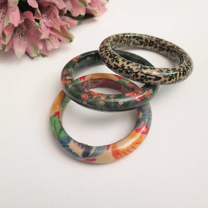 Puede incluir: Tres brazaletes de madera con diseños florales. El brazalete superior tiene un diseño floral en blanco y negro, el brazalete del medio tiene un diseño floral en verde y rojo, y el brazalete inferior tiene un diseño floral en azul y naranja.