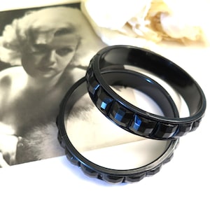 Könnte beinhalten: Zwei schwarze, facettierte, geometrische Armreifen liegen übereinander. Sie sind vor einer Vintage-Schwarz-Weiß-Fotografie einer Frau platziert. Die Armreifen haben ein glänzendes Finish.