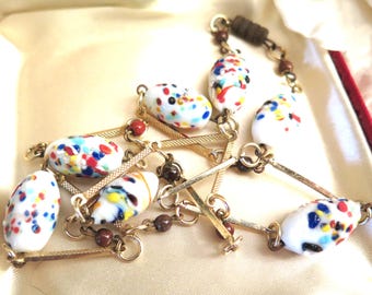 May include: A vintage necklace with white, oval beads adorned with colourful speckles. The beads are linked by gold-tone metal bars and small brown beads. The necklace is presented in a satin-lined box, highlighting its classic design.
