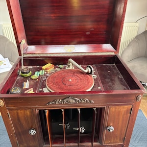 May include: A dark wood cabinet phonograph with a hinged lid, featuring a red turntable platter and a silver tone arm. The lower section includes two doors with decorative knobs and an open central storage compartment with vertical dividers.