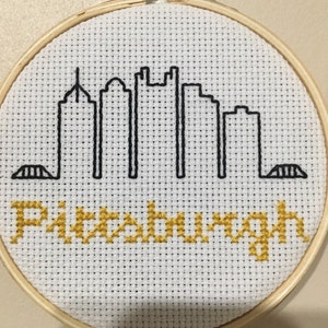 May include: A white embroidery hoop with a black and white cross-stitch design of the Pittsburgh skyline. The word "Pittsburgh" is stitched in gold below the skyline.