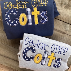 May include: Two sweatshirts with the text "Cedar Cliff Colts" embroidered on them. The text is in yellow with different patterned fabric accents. The top sweatshirt is navy blue and the bottom sweatshirt is gray.
