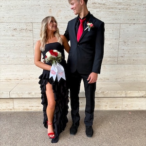 Könnte beinhalten: Ein Paar in festlicher Kleidung, die Frau in einem schwarzen, gerafften Kleid mit einem roten und weißen Blumenstrauß, der Mann in einem schwarzen Anzug mit einer roten Krawatte. Sie stehen vor einer weißen gekachelten Wand.