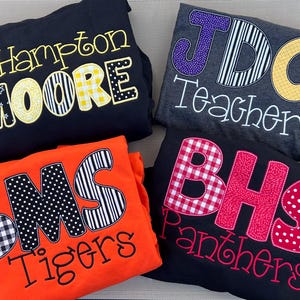 May include: Four folded sweatshirts in black, dark gray, and orange. Each features large, multi-patterned fabric applique letters and embroidered text. The designs include J. Hampton Moore, JDC Teacher, BMS Tigers, and BHS Panthers in various colors.