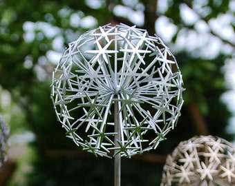 May include: Three silver metal spheres with a starburst design, mounted on tall metal poles. The spheres are arranged in a row, with the middle sphere in focus. The spheres are set against a blurred background of green foliage.