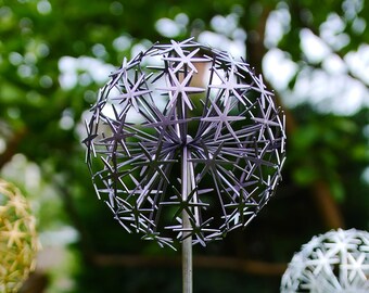 May include: Close-up of three metal dandelion sculptures in a garden setting. The central sculpture is silver, with a spherical head composed of interconnected star shapes. The other two are gold and white. The background is blurred green foliage.