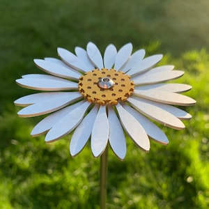May include: A white metal daisy with a gold centre and a silver screw in the centre. The daisy is on a metal stem and is standing in a grassy area.