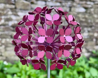 May include: A purple metal flower with a sphere shape and multiple petals. The flower is on a metal stem and is in front of a green leafy background.